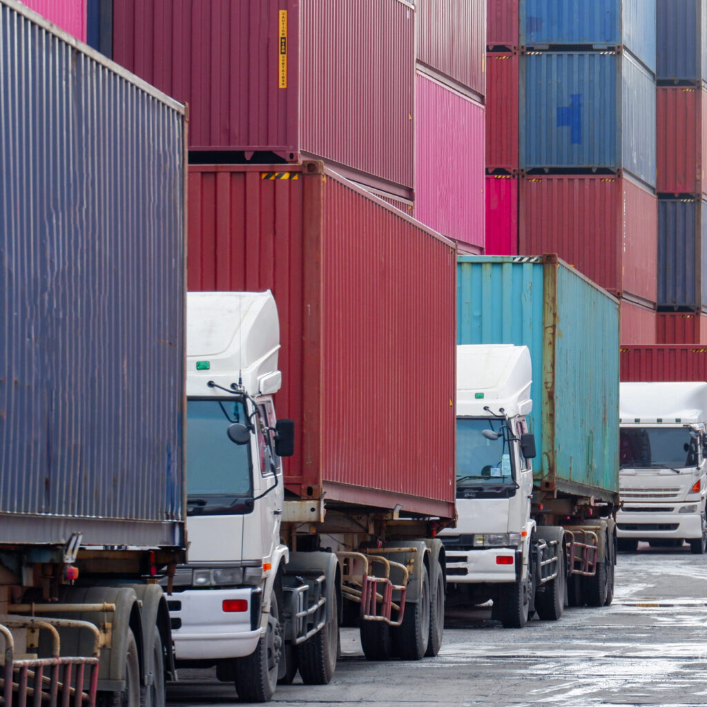 Image of a Logistics Yard with Container stacks in the background and a line of trucks leaving in the foreground.